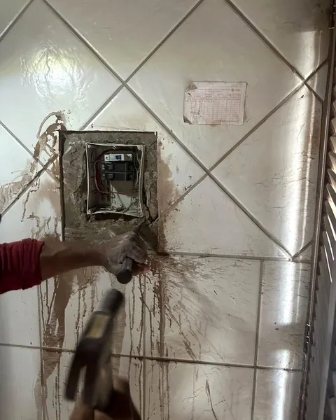 Parede de azulejo sendo aberta a marteladas, com o buraco onde vai o quadro de distribuição