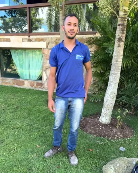 Eduardo de pé no gramado de uma casa, de camisa azul e calça jeans